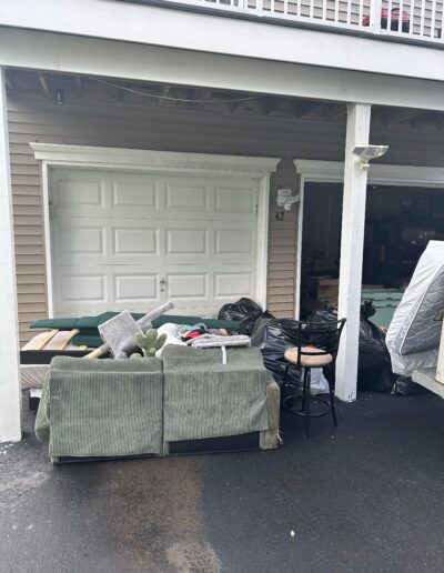 Cluttered driveway in front of a house with a green sofa, cushions and bags piled up in front of a closed garage door.