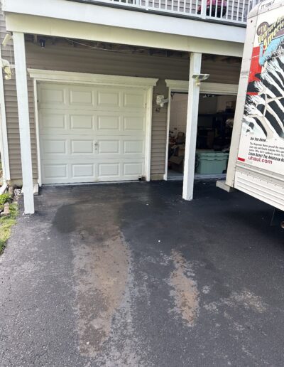 White sectional garage door under a raised deck, with a doorway and clutter visible to the right; a moving truck is parked adjacent to the driveway on the right side.