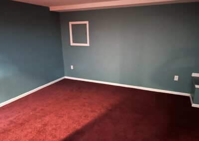 Empty room with teal walls, white baseboards, and red carpet; a small square-framed cabinet door mounted on the wall.