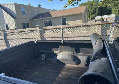 View from inside the bed of a dark dump truck, with dirt on the floor, a coiled hose, and a chain-link fence with houses in the background.