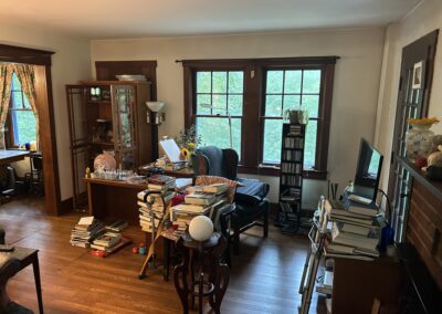 Cluttered living room with stacked books on tables and floor, wooden furniture, and large bay windows showing greenery outside.