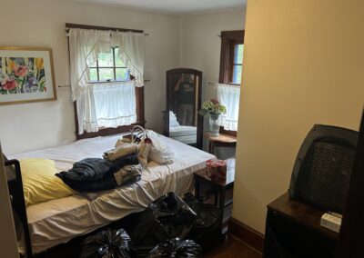 Messy bedroom with two beds, clothes scattered on the foreground, and several black trash bags on the floor; a mirror reflects a person taking the photo.