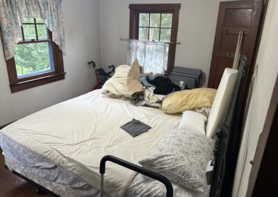 Messy bedroom with an unmade bed, piled laundry, and a walker near windows with floral curtains.