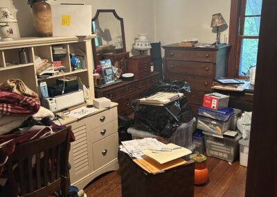 Cluttered bedroom or storage room with dressers, bins, papers, and clothes piled on furniture.