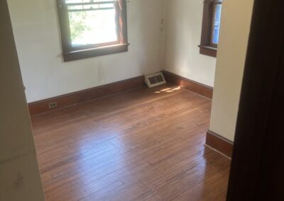 Small empty room with two wooden-trimmed windows, sunlight streaming in, and a floor vent near the baseboard.