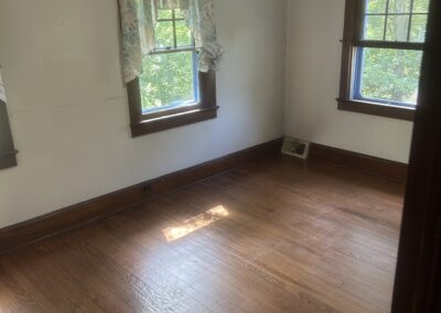 Empty room with hardwood floor, two windows, and floral curtains on the left window; ceiling light fixture overhead.