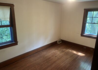 Empty beige-walled room with two wooden-framed windows, hardwood floor, and a floor vent near the baseboard.