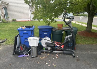 Cluttered front yard with blue recycling bins, a treadmill and assorted items next to the driveway under a tree in a suburban home settingicased by green lawn and a two-story house.