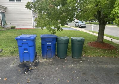 Four recycling/garbage bins in blue and green line the curb on a driveway in a suburban yard, with a large tree and a house to the left and a street in the background.